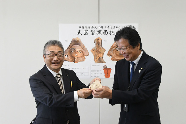 写真：副賞である表裏型顔面把手樹脂製模型を持つ西さん（左）と教育長（右）