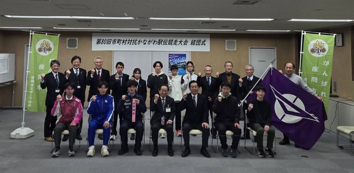 写真：「第80回市町村対抗かながわ駅伝競走大会結団式」と書かれた看板の下で、選手と出席者がガッツポーズで写真を撮っている様子